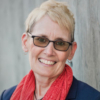 Photograph of Julie Derry, a woman with dark glasses, short blond hair, a red scarf, and a big smile.